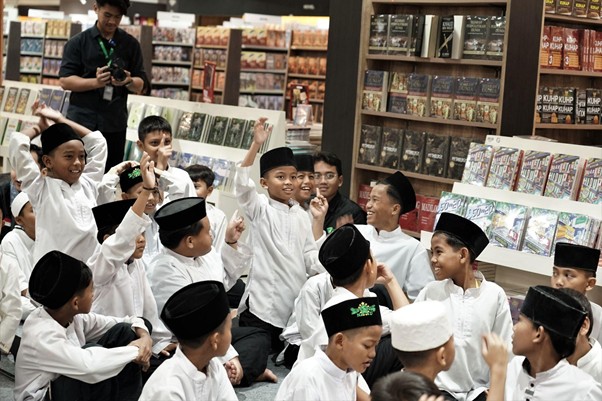 belanjabarengyatimparagon90 dompet dhuafa Group of children in white shirts and black caps gather in a bookstore, raising hands in celebration while a photographer watches in the background.