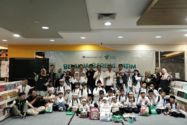 belanjabarengyatimparagon92 dompet dhuafa Large group of children and adults posing for a group photo indoors at an event, with backpacks on the floor and a banner in the background.