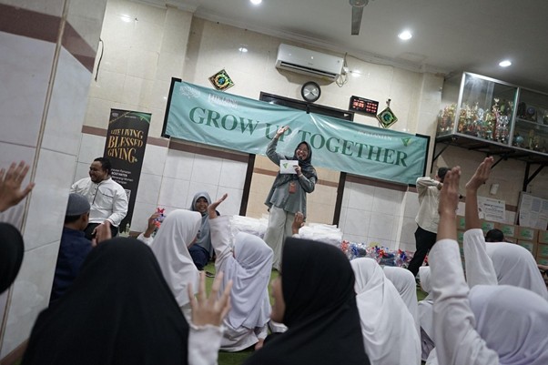 A speaker on stage addressing a crowd of students wearing white hijabs in a hall, many raising hands, with a banner reading 'GROW UP TOGETHER' behind them.