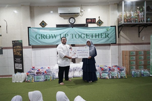 Man and woman present a certificate in a hall under a 'GROW UP TOGETHER' banner; donor bags and boxes line the wall, with an audience watching.