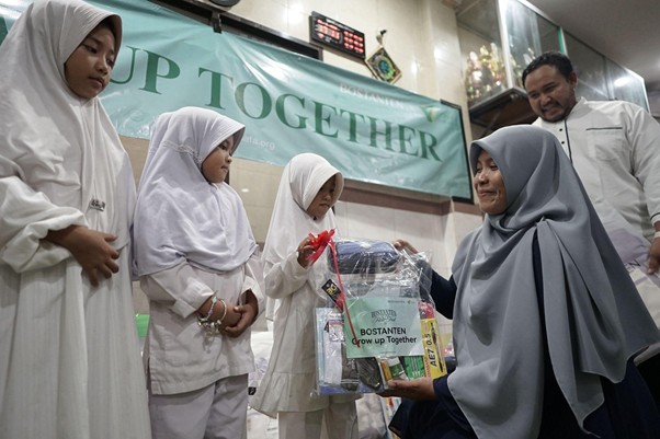 Woman in a gray hijab hands a gift pack to a girl in a white uniform at a 'Grow Up Together' event, with two other girls nearby.