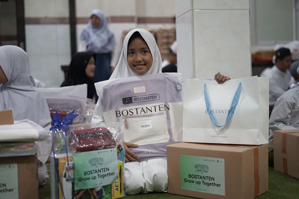 Young girl in a white hijab sits on the floor with gift bags and boxes labeled BOSTANTEN at a charity giveaway event.