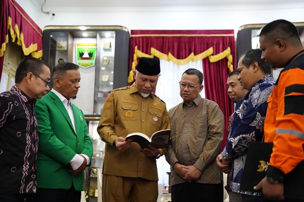 bukufotobingkas2 dompet dhuafa A group of men gathered around a man in a tan uniform who is reading from a book in a formal ceremony.
