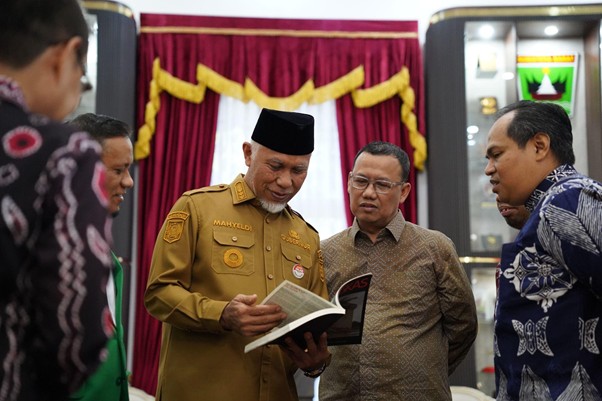 bukufotobingkas3 dompet dhuafa Man in a tan government uniform reads a booklet as two colleagues listen in a ceremonial setting with red curtains in the background.