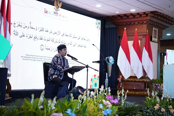 dampakzakatroisojkdandompetdhuafa20262 dompet dhuafa Man in a batik shirt sits at a microphone on a stage, reading from a music stand, with a large Arabic text screen behind and Indonesian flags nearby.