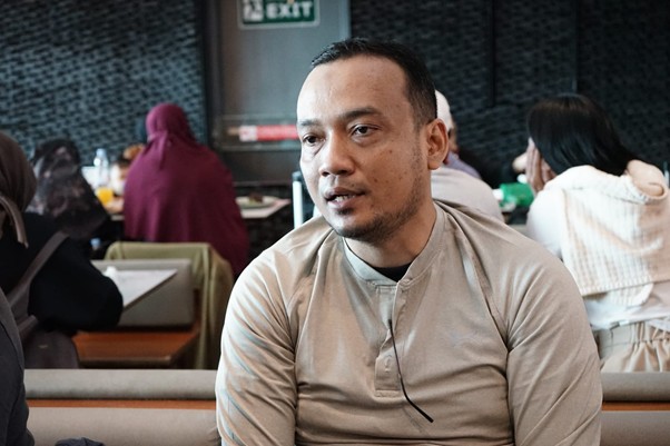delegasigpcidompetdhuafakebrusseluntukperdamaiandipalestina2 dompet dhuafa Man in a beige long-sleeve shirt sits in a cafe, talking, with other diners in the background.