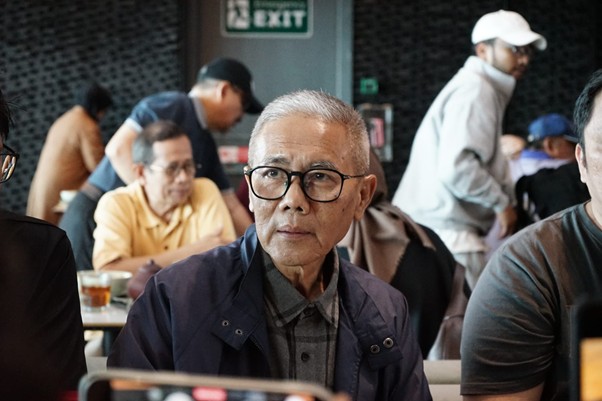 delegasigpcidompetdhuafakebrusseluntukperdamaiandipalestina5 dompet dhuafa Older man with gray hair and glasses seated at a cafe table, looking toward the camera; busy background with other diners.