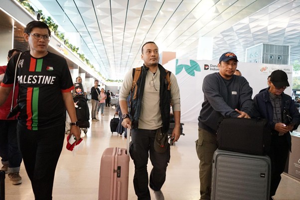 delegasigpcidompetdhuafakebrusseluntukperdamaiandipalestina dompet dhuafa Three men with rolling suitcases walk through a bright airport terminal; one wears a Palestine-themed shirt and backpack.