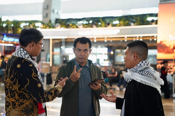 Three men in an airport concourse chat; the center man holds a phone and shows a peace sign as the others gesture.