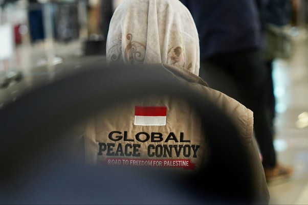 Back view of a person in a beige jacket with a 'GLOBAL PEACE CONVOY' logo and a red-and-white flag patch, text 'Road to Freedom for Palestine' beneath.