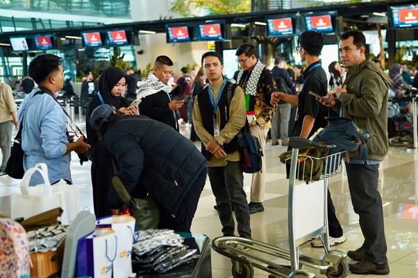 Group of travelers with luggage and a baggage cart waiting at an airport check-in area, some looking at their phones amidst displays overhead.