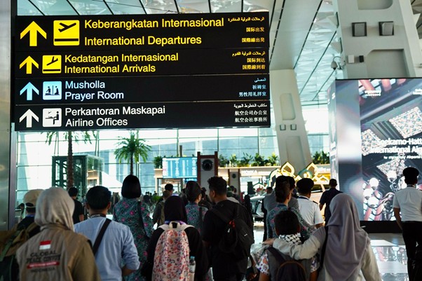 Airport signboard in a busy terminal shows international departures and arrivals, a prayer room, and airline offices, with travelers walking underneath.