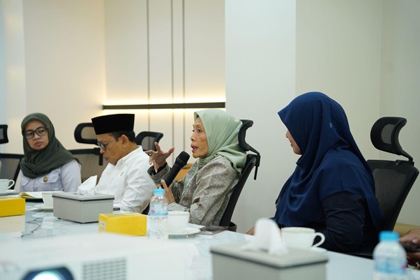 A woman in a light green hijab speaks into a microphone at a conference table, surrounded by colleagues in headscarves.