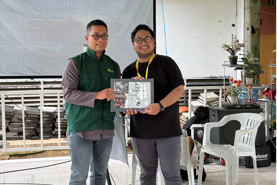 donasifiber1 dompet dhuafa Two men pose indoors, one in a green vest hands a framed award to the other, smiling at the camera.