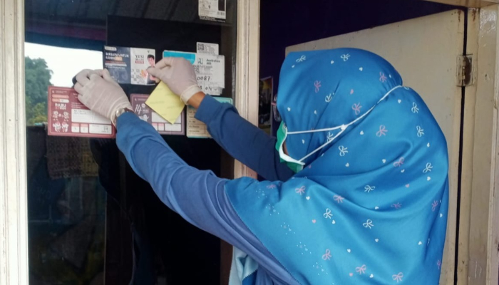 fotojet3 dompet dhuafa Woman in a blue patterned hijab and mask posts a yellow notice on a shop's bulletin board by the doorway, wearing gloves.