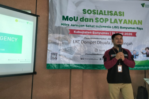 A man in a dark vest speaks into a microphone beside a banner about MoU and SOP layanan at a seminar; a projector screen on the left shows 'EMERGENCY'.