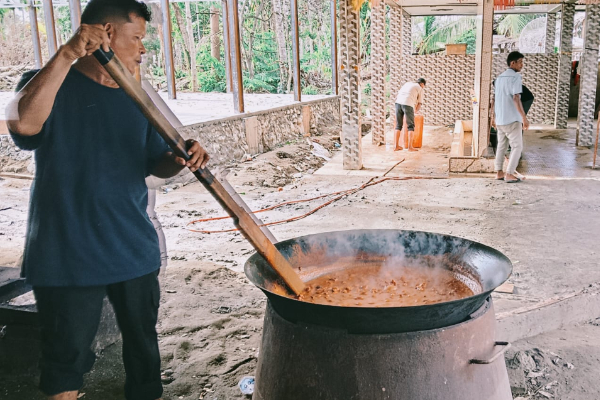 Man stirs a large pot over an open fire with a long wooden paddle in a partially built outdoor kitchen; others work in the background.