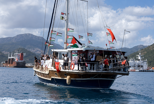 A wooden sailing yacht crowded with passengers on deck, decorated with pennant flags, cruising in calm blue water with mountains in the background.