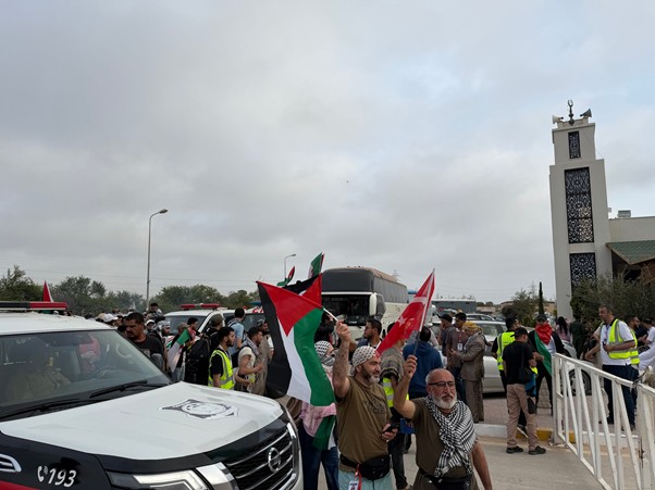 global sumud flotilla afrika utara 1 dompet dhuafa Crowd of protesters waving Palestinian flags near a bus and security personnel at a public square.