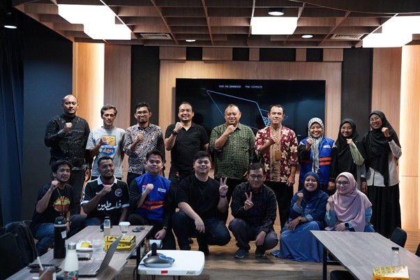Group of 16 people posing for a photo in a modern conference room with a large screen in the background, some giving thumbs up or peace signs.
