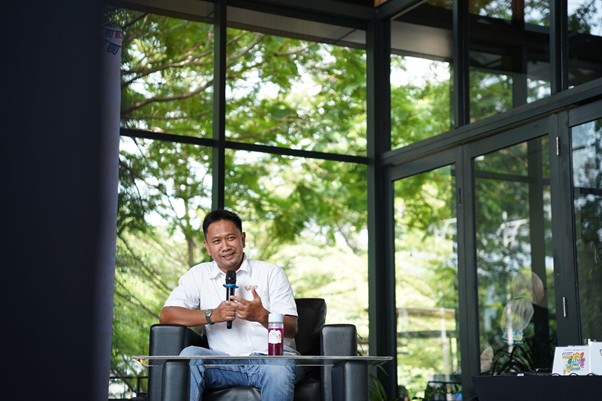greenjobsdanekonomihijau2 dompet dhuafa Man in a white shirt seated on a black chair, speaking into a microphone in a glass-walled room with greenery outside.