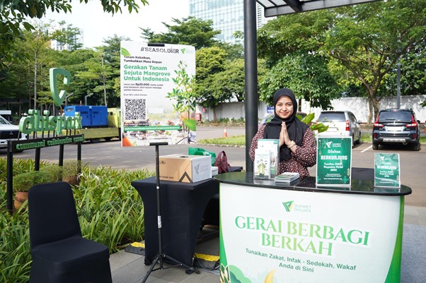 greenjobsdanekonomihijau7 dompet dhuafa Smiling woman in a hijab runs a charity booth labeled 'Gerai Berbagi Berkah' outdoors, with donation boxes and brochures for zakat, infaq, sedekah, and wakaf nearby.