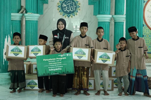 Group of children and a woman in a mosque, posing with donor boxes for a distribution program in front of teal pillars.
