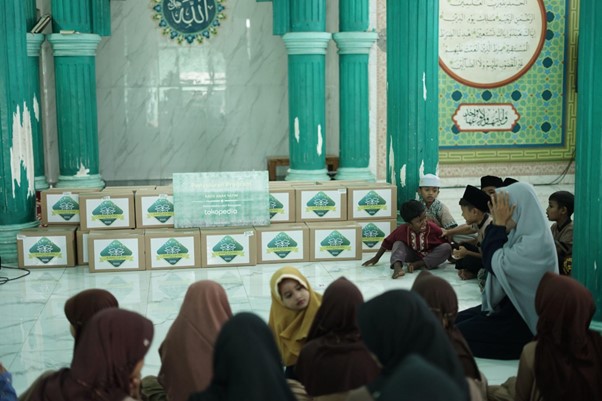 Group of people, mostly women and children, seated on a mosque floor facing a stack of labeled donation boxes in a teal-colored hall.