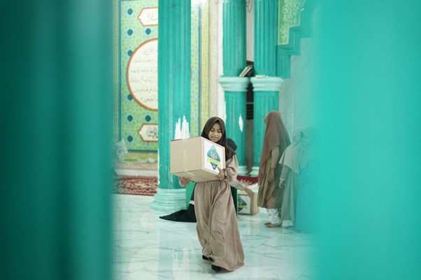 Young woman in a brown abaya carrying a cardboard box through turquoise columns in a mosque-like hallway.
