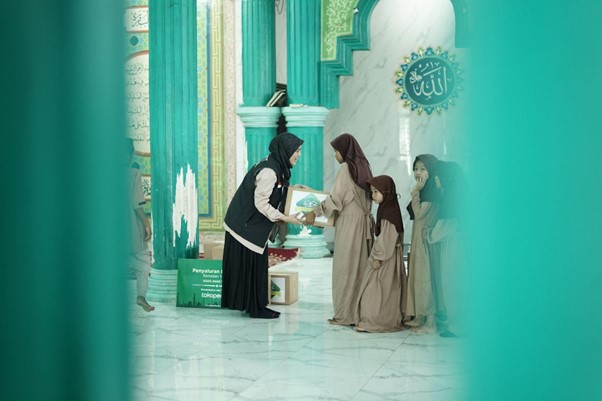 A woman wearing a black headscarf and vest hands a pamphlet to a girl in beige robes inside a mosque with green columns, while two others watch.