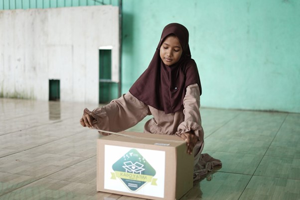 Girl in a brown hijab places a ballot into a cardboard voting box on a tiled floor.