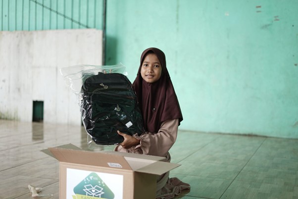 Girl wearing a maroon hijab kneels beside an open donation box, holding a new black backpack wrapped in plastic in a bare, green-walled room.