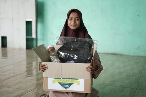 Smiling girl in a hijab holds a cardboard donation box containing a backpack and other items.