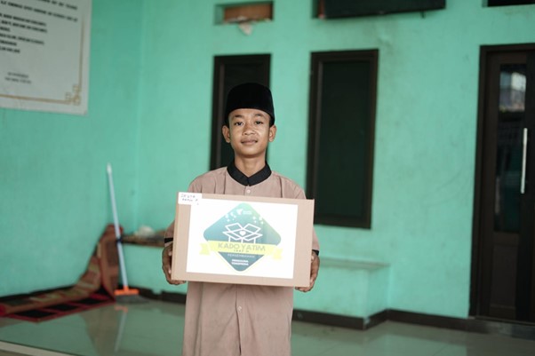 Boy in traditional beige outfit and black cap holding a cardboard box with a charity logo, indoors against a turquoise wall.