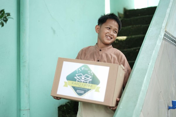 Smiling boy in a brown traditional shirt carrying a cardboard box with a green charity logo outside near a green stairwell.