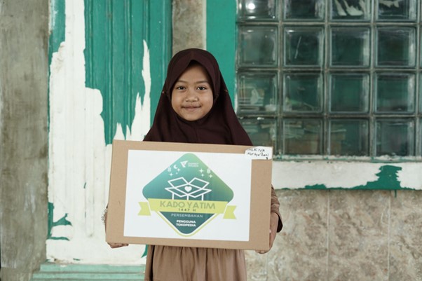 Girl in a brown hijab and dress holding a cardboard sign with a Kado Yatim charity logo, standing outside a green-painted wall and glass block window.
