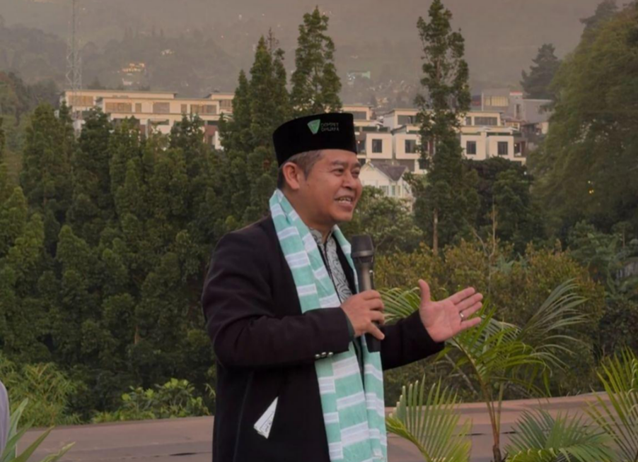 hotelariandripuncak4 dompet dhuafa Man in a black traditional hat and green-striped scarf speaks into a handheld microphone outdoors, gesturing with his other hand, with trees and buildings in the background.