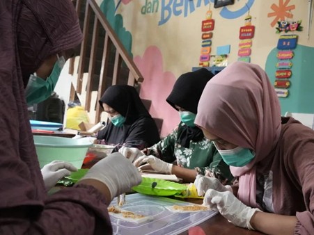 Women in hijabs and masks wearing gloves, seated at a table and assembling items in a classroom workshop setting.