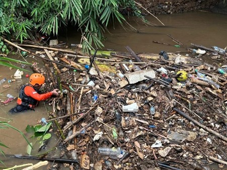 Person in an orange helmet wades in muddy water along a trash-strewn riverbank, cleaning up debris like plastic and wood.