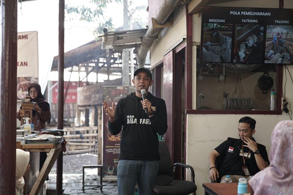 jelajahkandanguntukkurban202613 dompet dhuafa Man in a black shirt speaks into a microphone to a small outdoor audience at a farm/market booth.