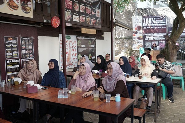 jelajahkandanguntukkurban202614 dompet dhuafa Group of mostly women wearing hijabs sit at outdoor wooden tables at a halal food stall, with menus and posters on the storefront behind them.