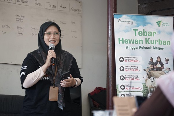jelajahkandanguntukkurban20262 dompet dhuafa Woman in a hijab speaks into a microphone at a presentation, holding a phone, with a whiteboard and a 'Tebar Hewan Kurban' poster beside her.