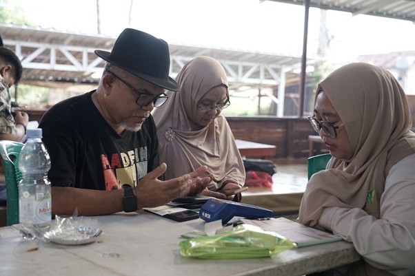 jelajahkandanguntukkurban20267 dompet dhuafa Three people sit at a table in a workshop, the man in a hat showing something on his hands to two women wearing hijabs.
