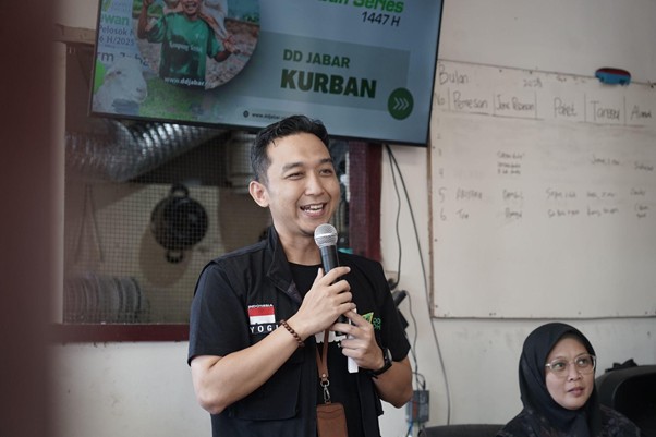 jelajahkandanguntukkurban20268 dompet dhuafa Man speaking into a microphone and smiling during a presentation, with a screen and whiteboard behind him and a woman in a hijab seated nearby.
