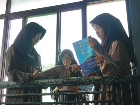 Three hijab-wearing students standing by a railing, reading and discussing books together outdoors by windows.