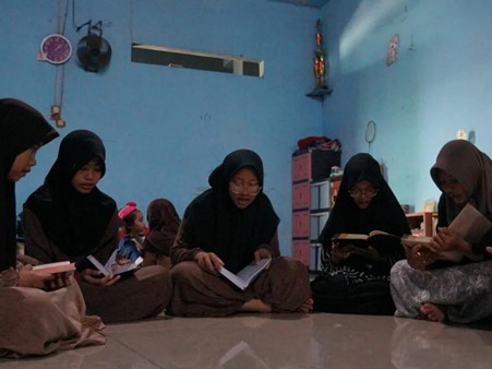 Group of women in hijabs sitting on the floor in a circle reading books in a blue classroom.