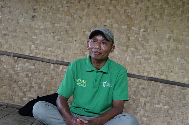 Man wearing a green polo shirt with logos sits cross-legged against a woven bamboo wall.