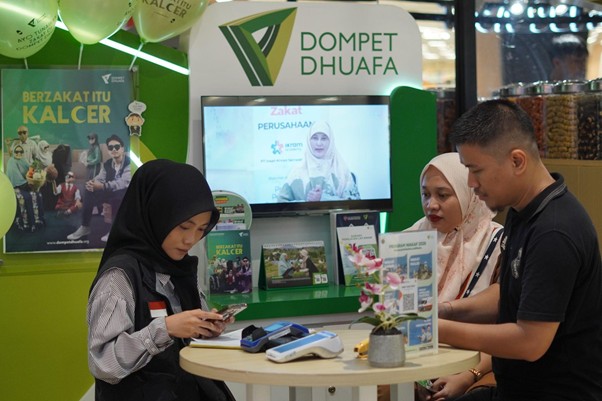 Three visitors at a Dompet Dhuafa booth: a woman in a black hijab uses her phone while a man and another woman look on near a round table and display screen.
