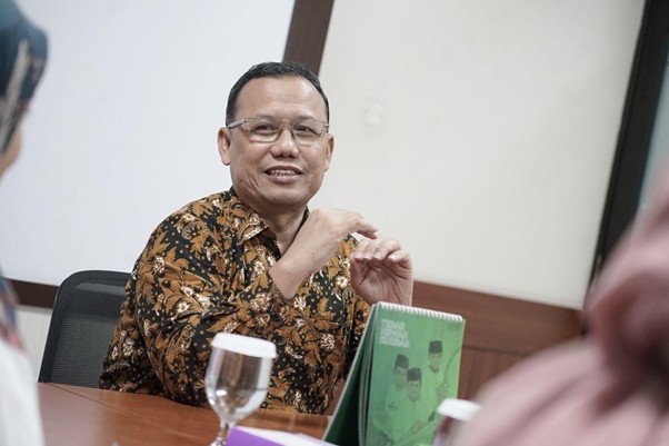 kolaborasimarcellazalianty2 dompet dhuafa Smiling man in a batik shirt sits at a conference table, gesturing with his hands during a meeting, with a green book and water glass nearby in an office setting.