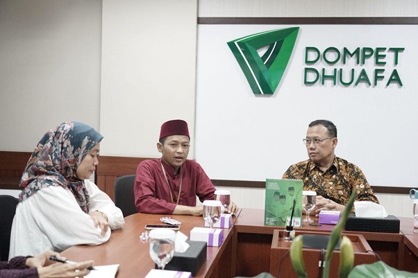 kolaborasimarcellazalianty3 dompet dhuafa Three people sit around a conference table in a meeting room, with a Dompet Dhuafa sign and green logo on the wall; the man in a maroon cap speaks while others listen.